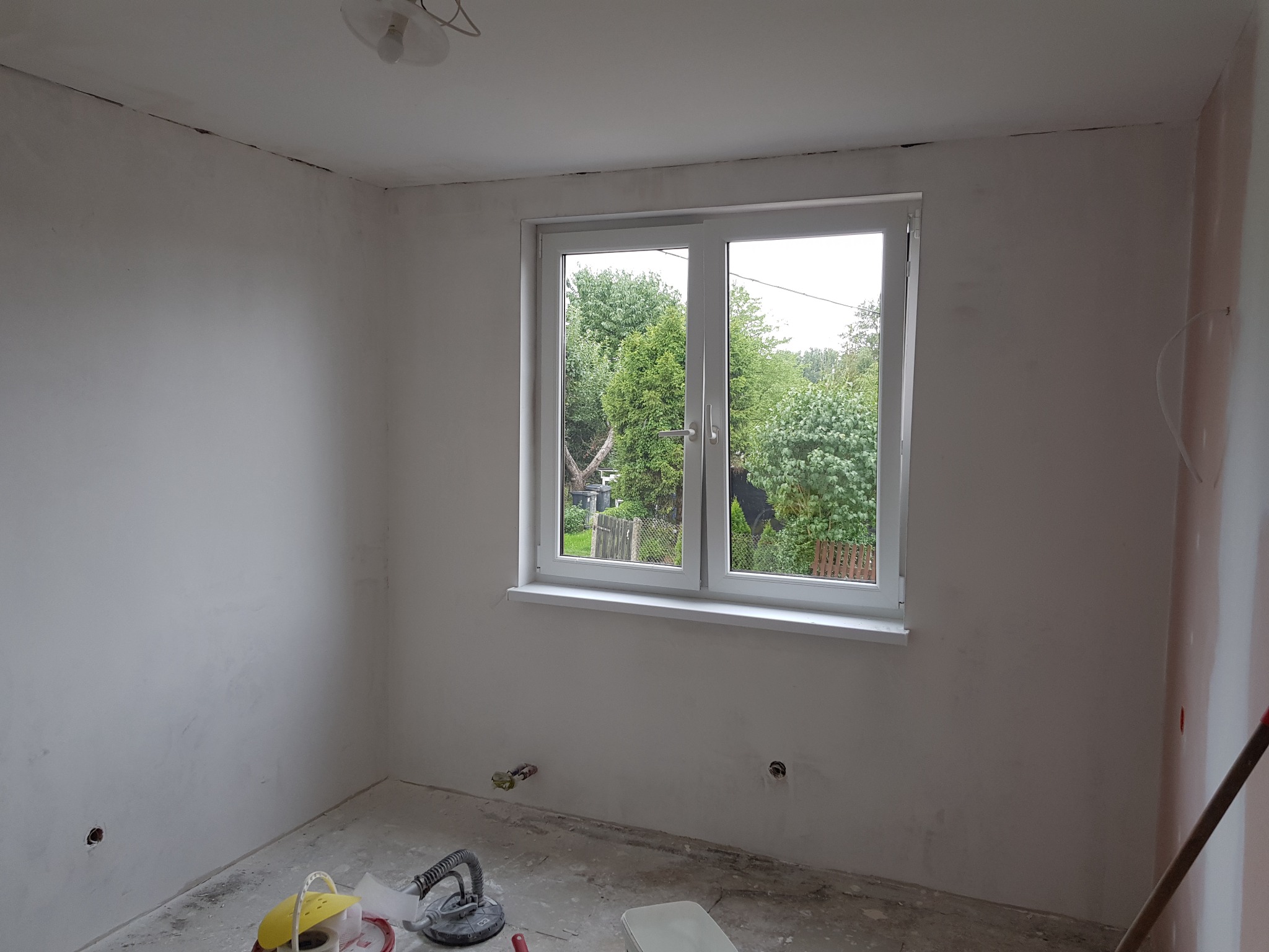 Pokój w trakcie przygotowania do malowania: ściany świeżo wyszpachlowane, widoczne narzędzia szlifierskie na podłodze, okno z widokiem na zieleń.