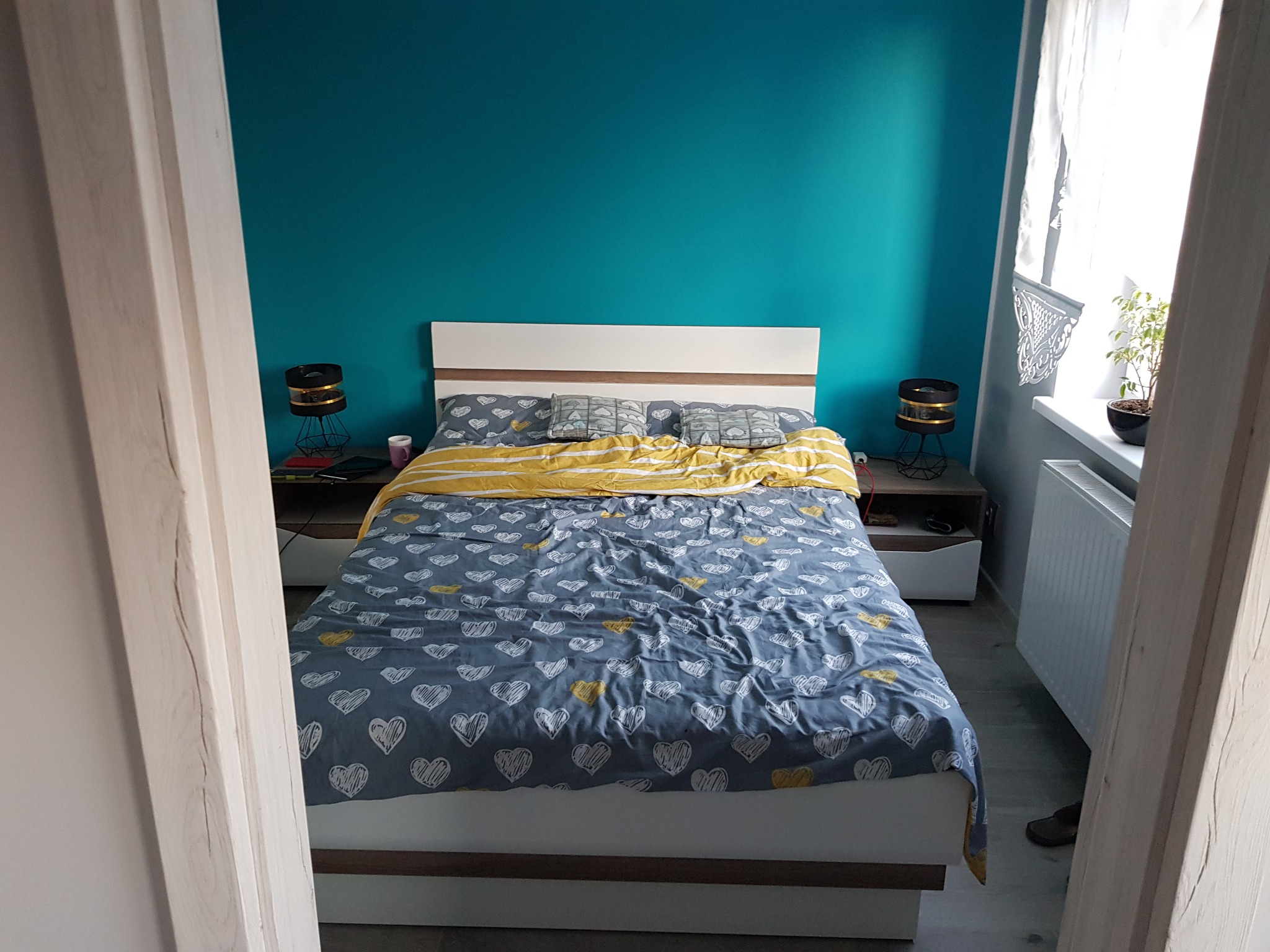 Sypialnia z łóżkiem w pościeli w szare serca na tle turkusowej ściany, widok przez framugę drzwi.