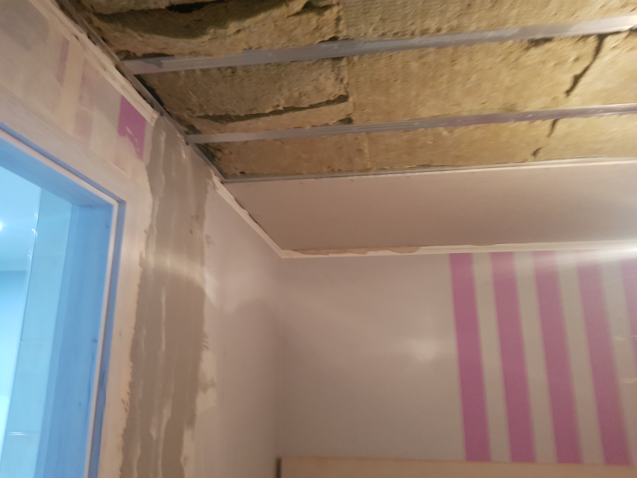 Poddasze w trakcie ocieplania z widoczną wełną mineralną, metalowymi profilami i częściowo zamontowaną płytą gipsowo-kartonową; fragment ściany z różowymi pasami w tle.