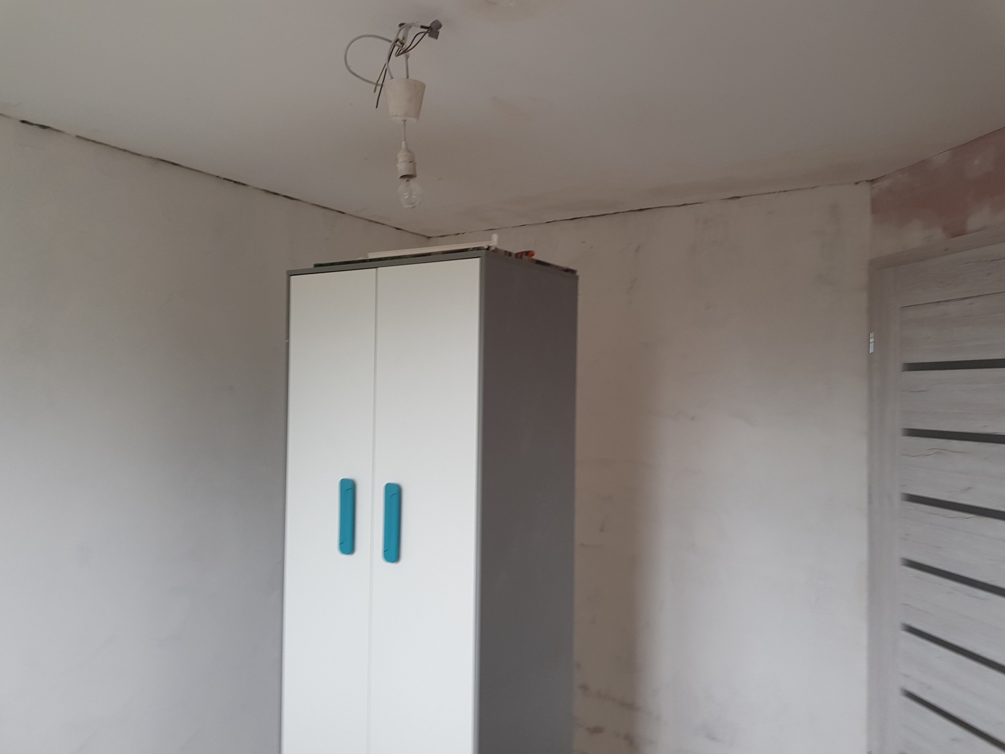 Pokój w trakcie prac wykończeniowych: surowe ściany, widoczne ślady po szpachlowaniu, wisząca żarówka na kablu oraz szafa z niebieskimi uchwytami.