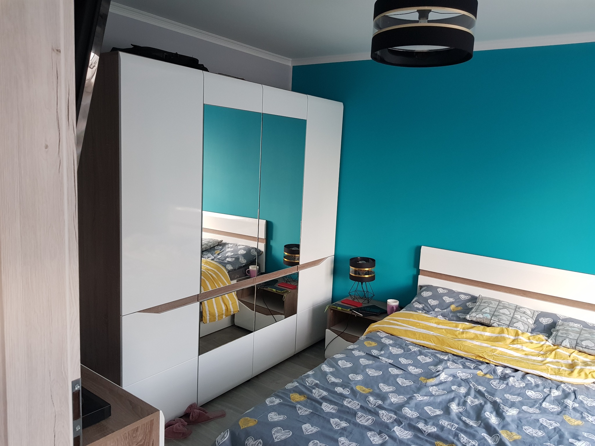 Nowoczesna sypialnia z turkusową ścianą, białą szafą z lustrami i łóżkiem w serduszka, widziana z boku.