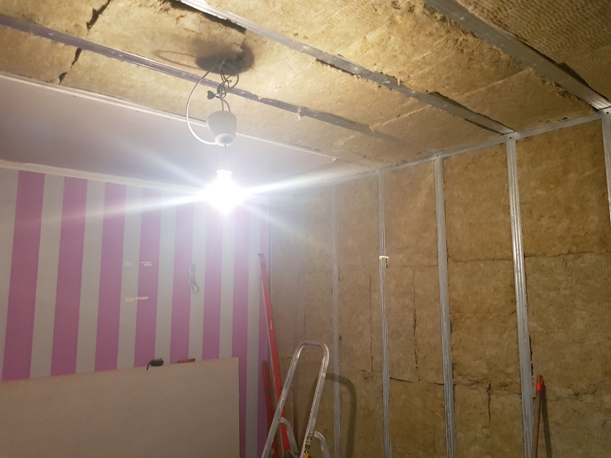 Poddasze w trakcie ocieplania wełną mineralną, widoczne metalowe profile konstrukcyjne, drabina oparta o ścianę w pasy, tymczasowe oświetlenie.