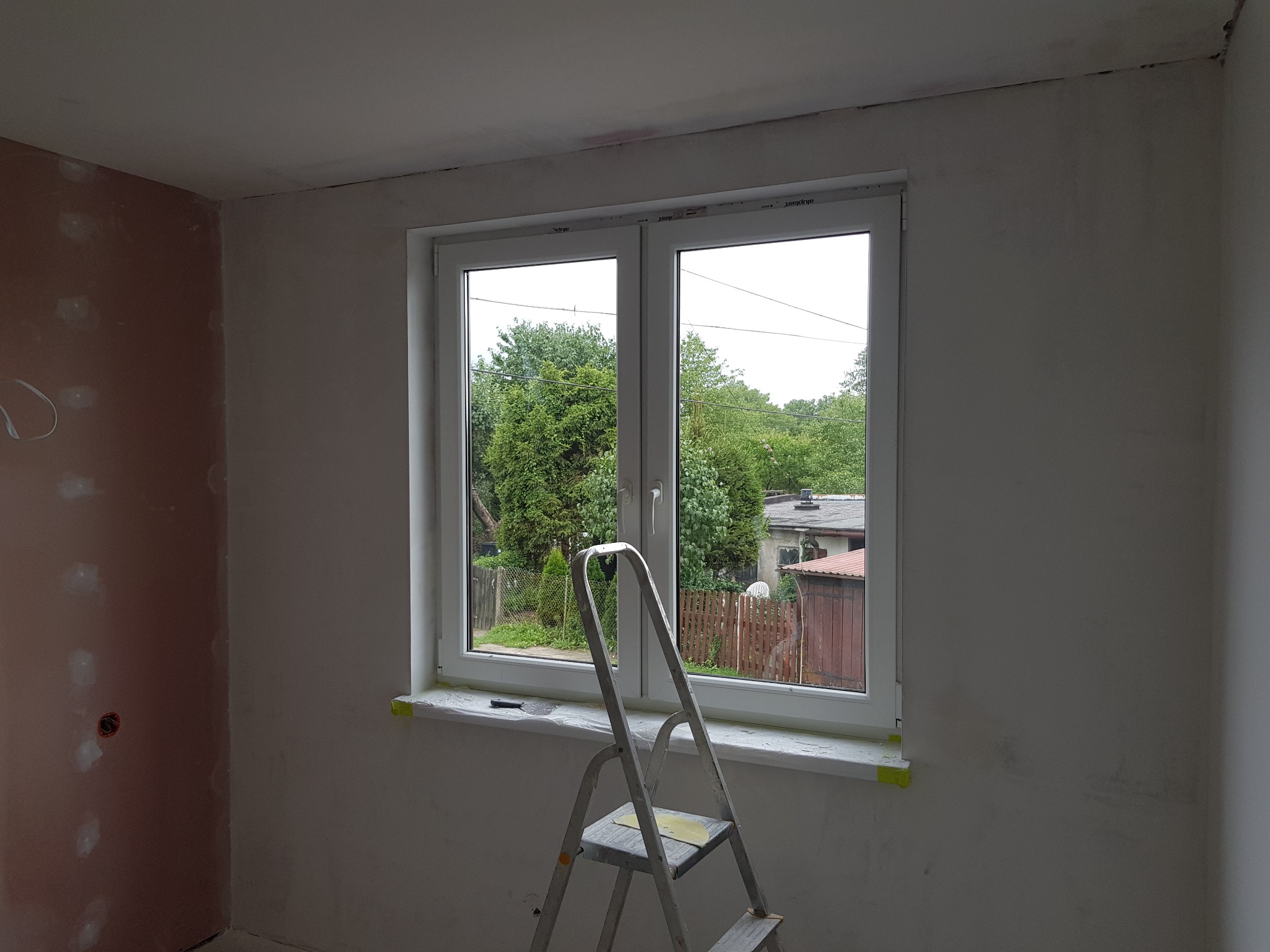 Pokój w trakcie przygotowania do malowania: ściany częściowo zagruntowane, okno zabezpieczone taśmą malarską, drabina aluminiowa oparta o parapet, widok na zieleń za oknem.