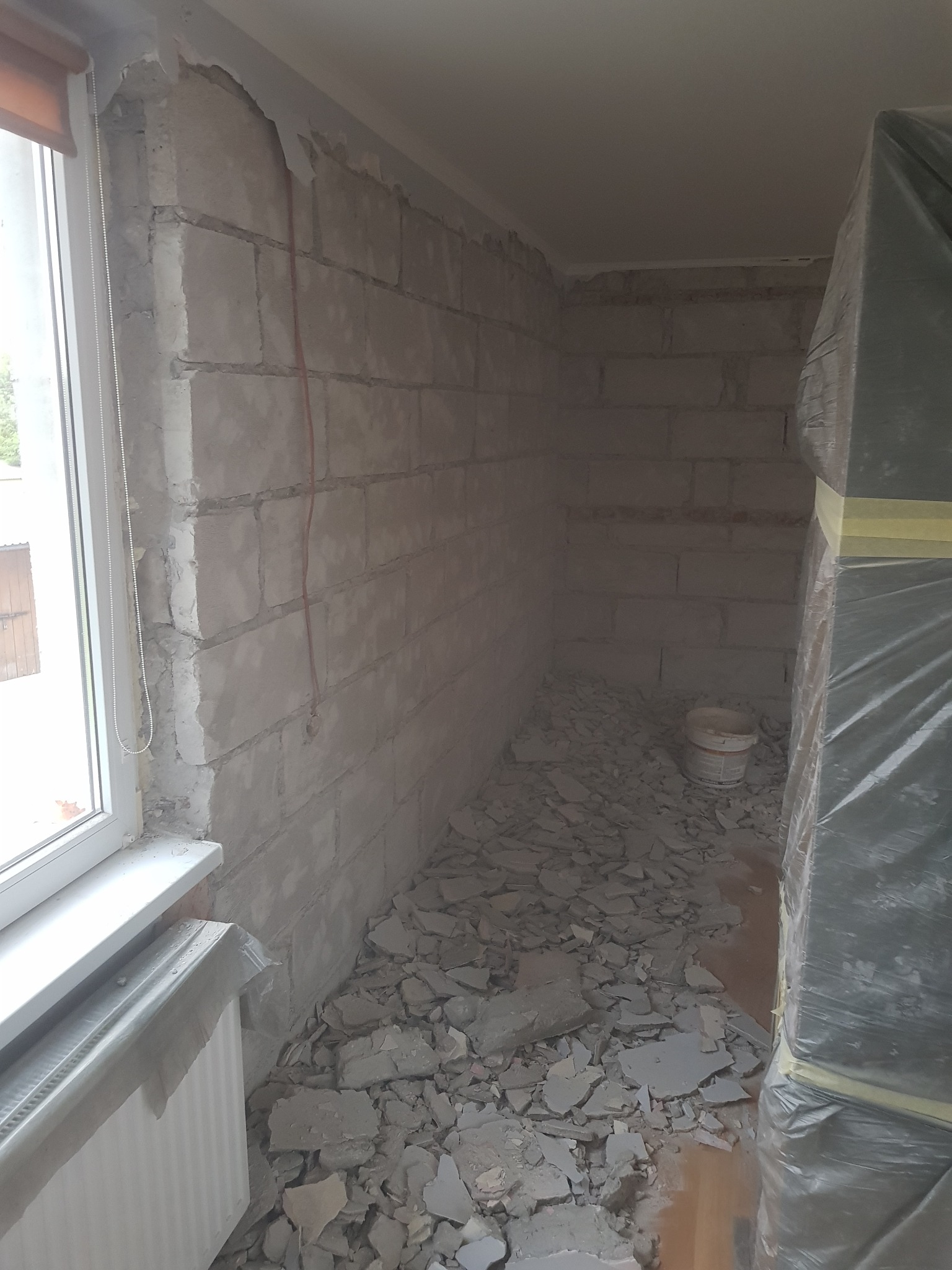 Wnętrze w trakcie prac demontażowych, widoczne ściany z betonu komórkowego, gruz na podłodze, zabezpieczony folią mebel i wiadro po farbie.