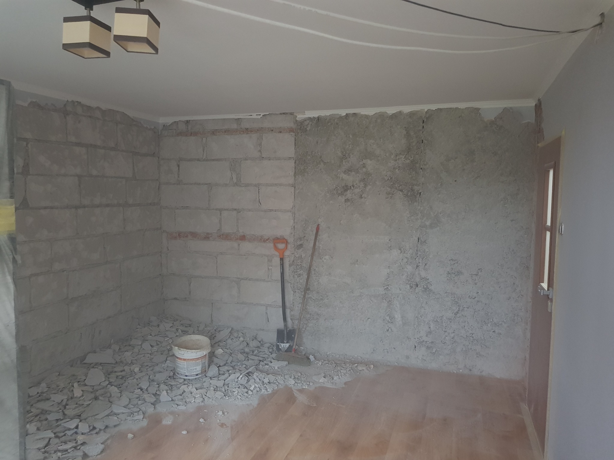 Pokój w trakcie prac przygotowawczych: ściany częściowo z surowego betonu komórkowego, częściowo otynkowane, gruz na podłodze, wiadro z zaprawą, łopata i miotła oparte o ścianę.