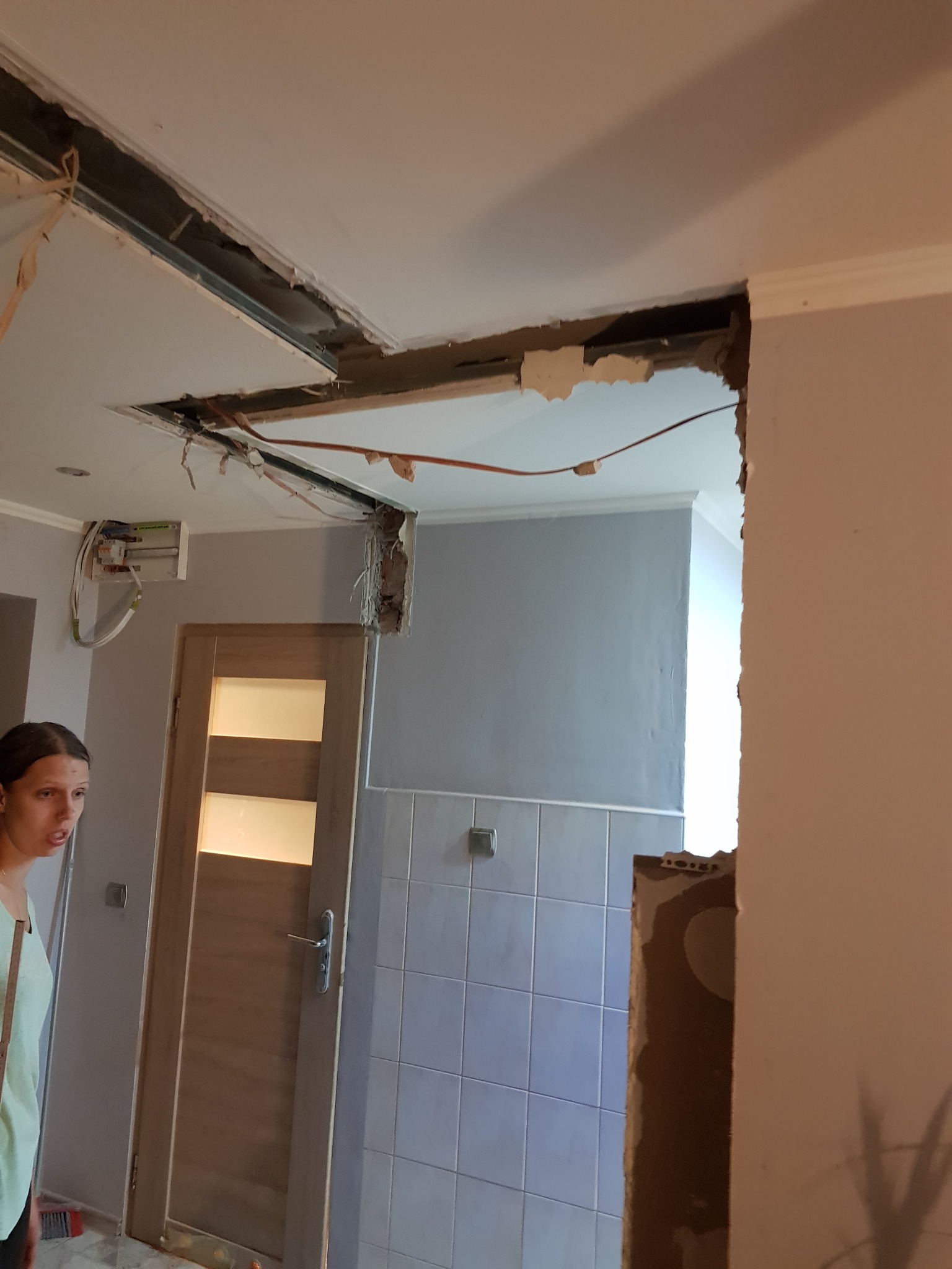 Uszkodzony sufit podczas prac, widoczne odsłonięte przewody i konstrukcja nośna, fragment ściany z ubytkami tynku, w tle drzwi z przeszkleniem i kobieta obserwująca prace.