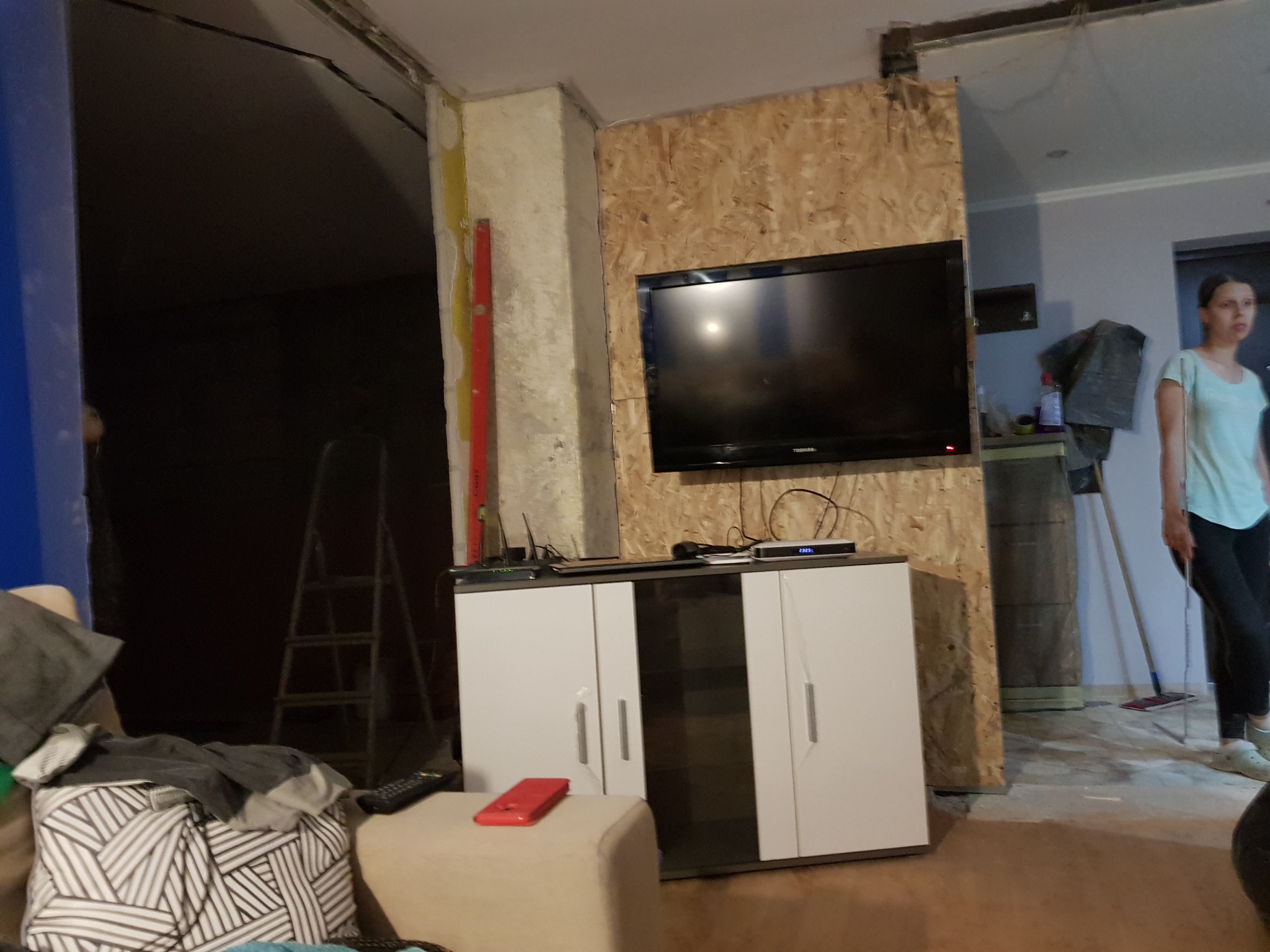 Wnętrze w Janowicach w trakcie prac wykończeniowych: widoczna ściana wyłożona płytą OSB, biała szafka RTV, drabina w tle i osoba sprzątająca podłogę.