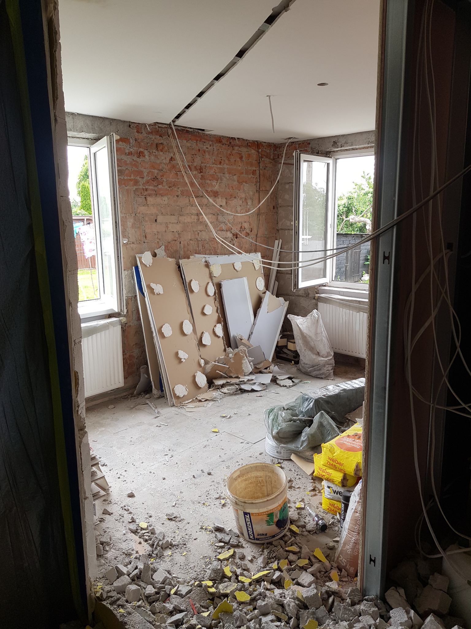 Pokój w trakcie remontu z odsłoniętą cegłą, częściowo wykończonym sufitem, widocznymi kablami, workami z zaprawą i gruzem na podłodze, oraz płytami gipsowymi opartymi o ścianę.