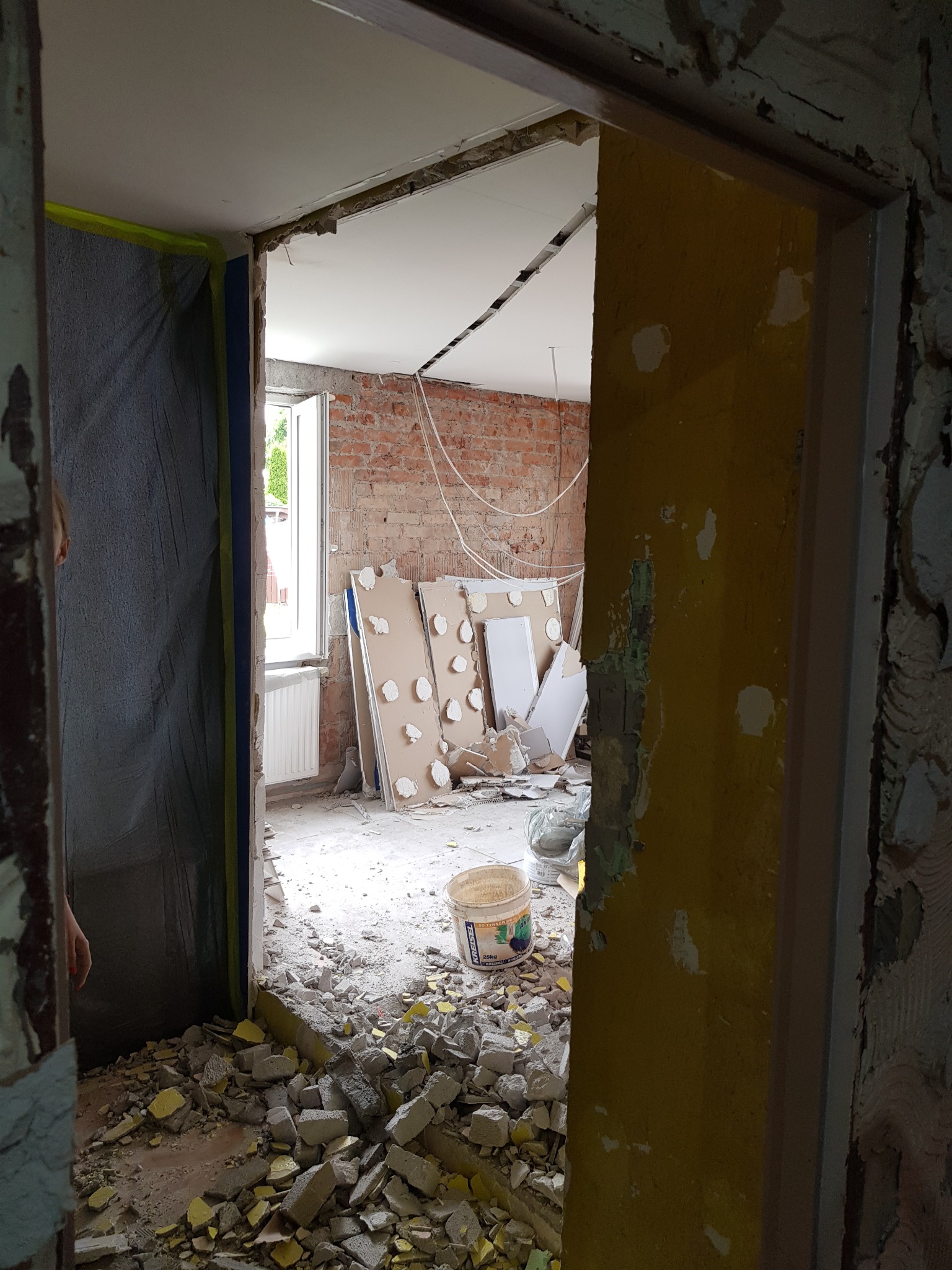 Wnętrze w trakcie demontażu: gruz na podłodze, odsłonięta cegła, płyty gipsowe oparte o ścianę, widok przez futrynę drzwiową, zabezpieczona folią ściana po lewej stronie.
