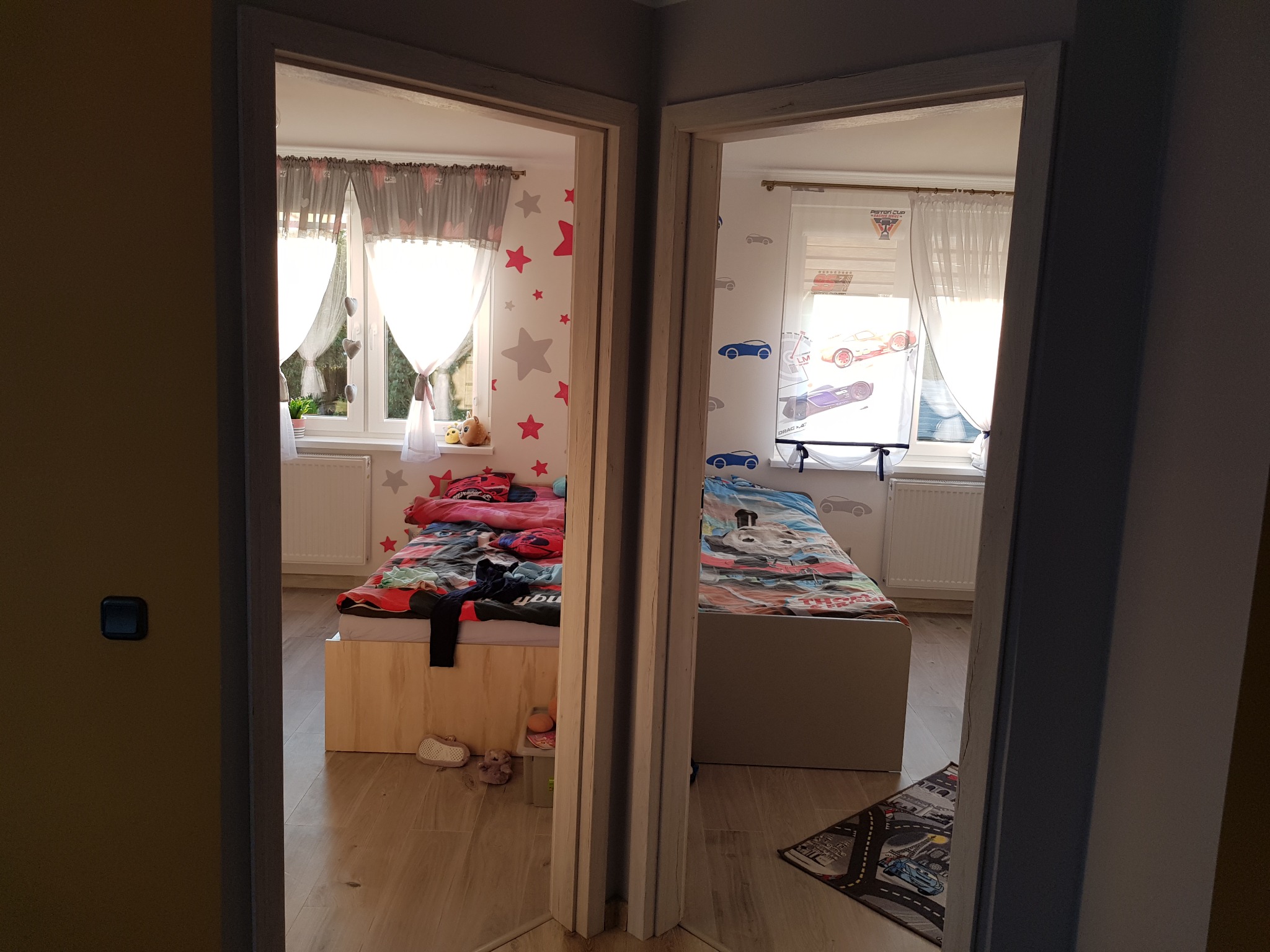 Widok przez podwójne drzwi do dwóch pokojów dziecięcych z łóżkami, oknami z zasłonami i dywanem z motywem ulic.