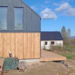 HARDYKAM Roman Olszyński - Nowoczesny dom szkieletowy z drewnianą elewacją i przyległym tarasem w trakcie budowy, widoczne materiały budowlane i niezagospodarowany teren.