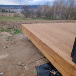 HARDYKAM Roman Olszyński - Nowo wybudowany taras z jasnego drewna z widokiem na krajobraz z drzewami i wieżą telekomunikacyjną w tle, widoczne elementy konstrukcyjne i czarna folia pod spodem.