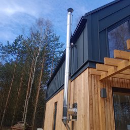 HARDYKAM Roman Olszyński - Zewnętrzny widok na fragment domu z drewnianą elewacją, ciemną blachą na dachu i kominem ze stali nierdzewnej, z widocznym stosem drewna w tle.