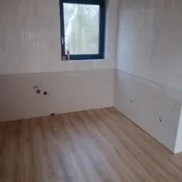 Łazienka w trakcie wykończenia: ułożone panele podłogowe, częściowo wyłożone płytki na ścianach, przygotowane przyłącza wodne i kanalizacyjne, okno z widokiem na zewnątrz.