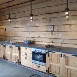 Rustykalna kuchnia w domku z bali, z jasnymi drewnianymi szafkami zdobionymi czarnymi okuciami, szarym blatem, wiszącymi lampami na sznurach i elementami z surowego drewna.