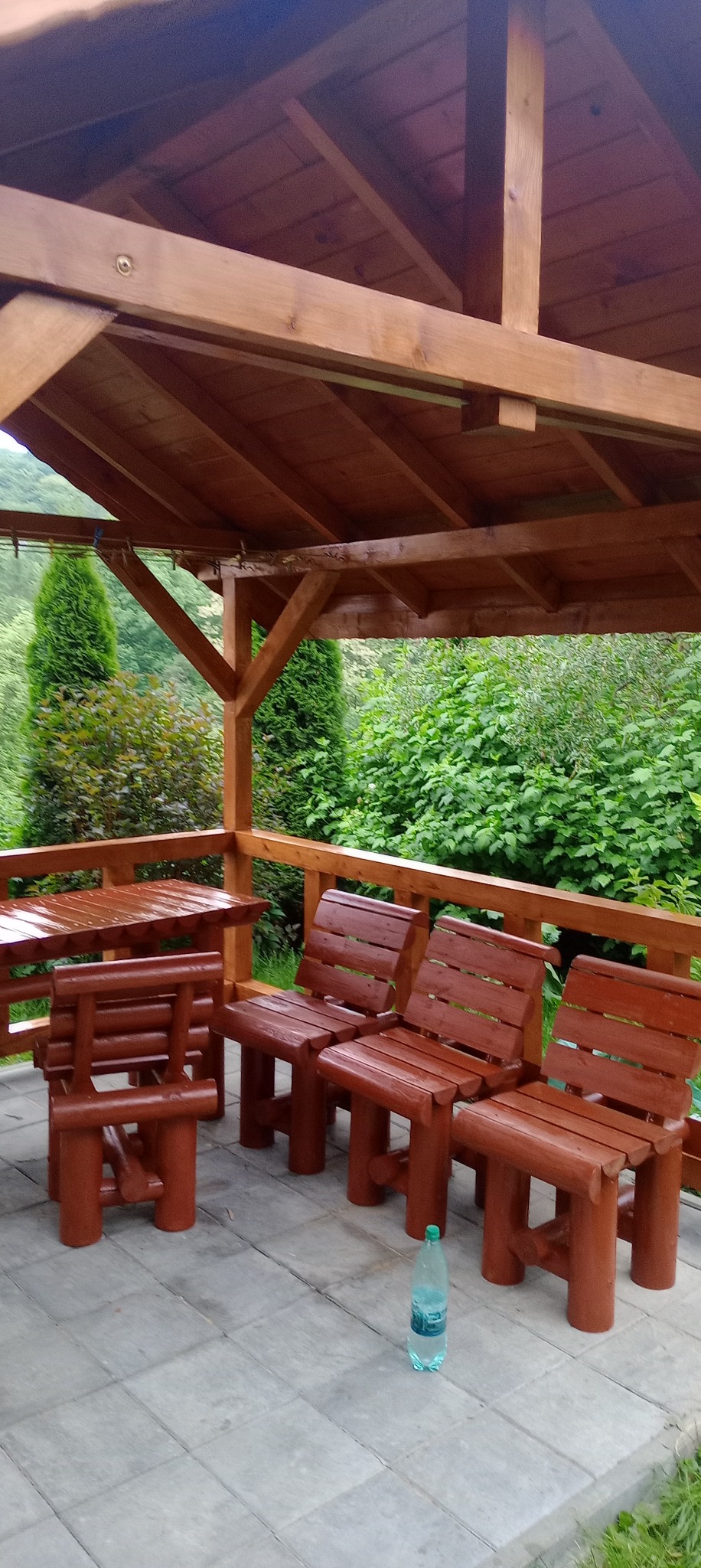 Zadaszony taras z drewnianą konstrukcją i meblami ogrodowymi w rustykalnym stylu, widok na zieleń, butelka wody na posadzce.