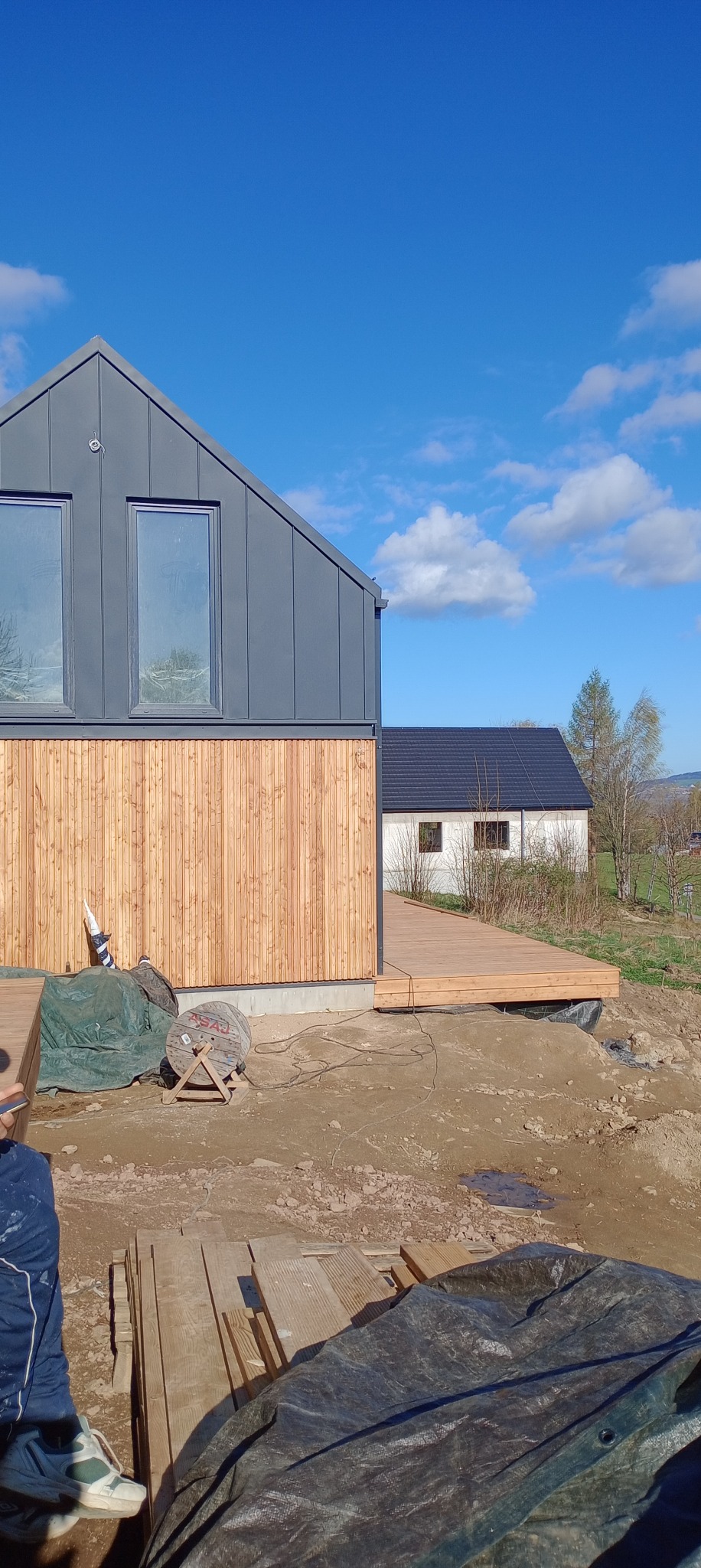 Nowoczesny dom szkieletowy z drewnianą elewacją i przyległym tarasem w trakcie budowy, widoczne materiały budowlane i niezagospodarowany teren.