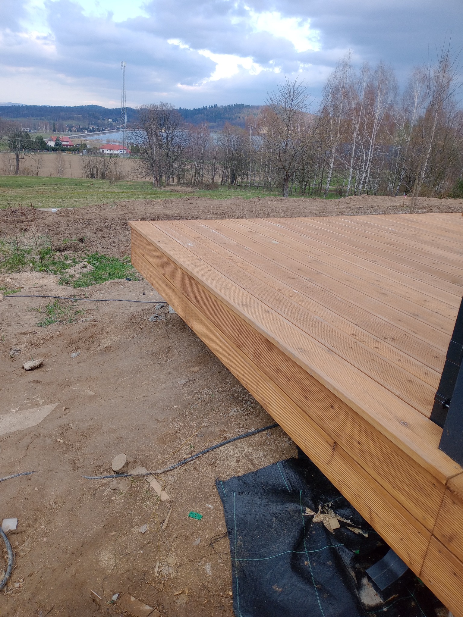 Nowo wybudowany taras z jasnego drewna z widokiem na krajobraz z drzewami i wieżą telekomunikacyjną w tle, widoczne elementy konstrukcyjne i czarna folia pod spodem.