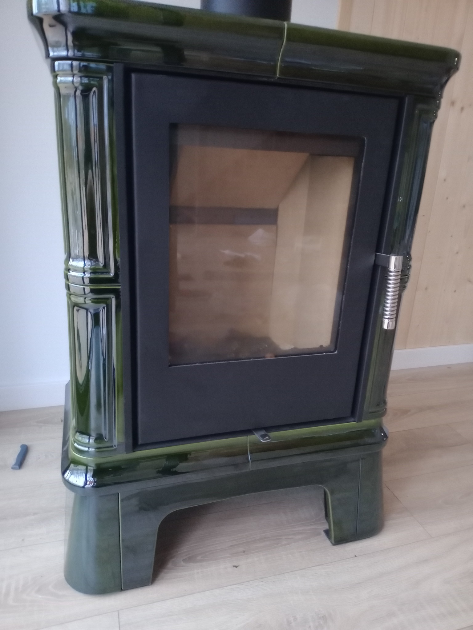 Glazowany, zielony piec kaflowy z czarnymi drzwiczkami i chromowanym uchwytem na tle drewnianej ściany i jasnej podłogi.