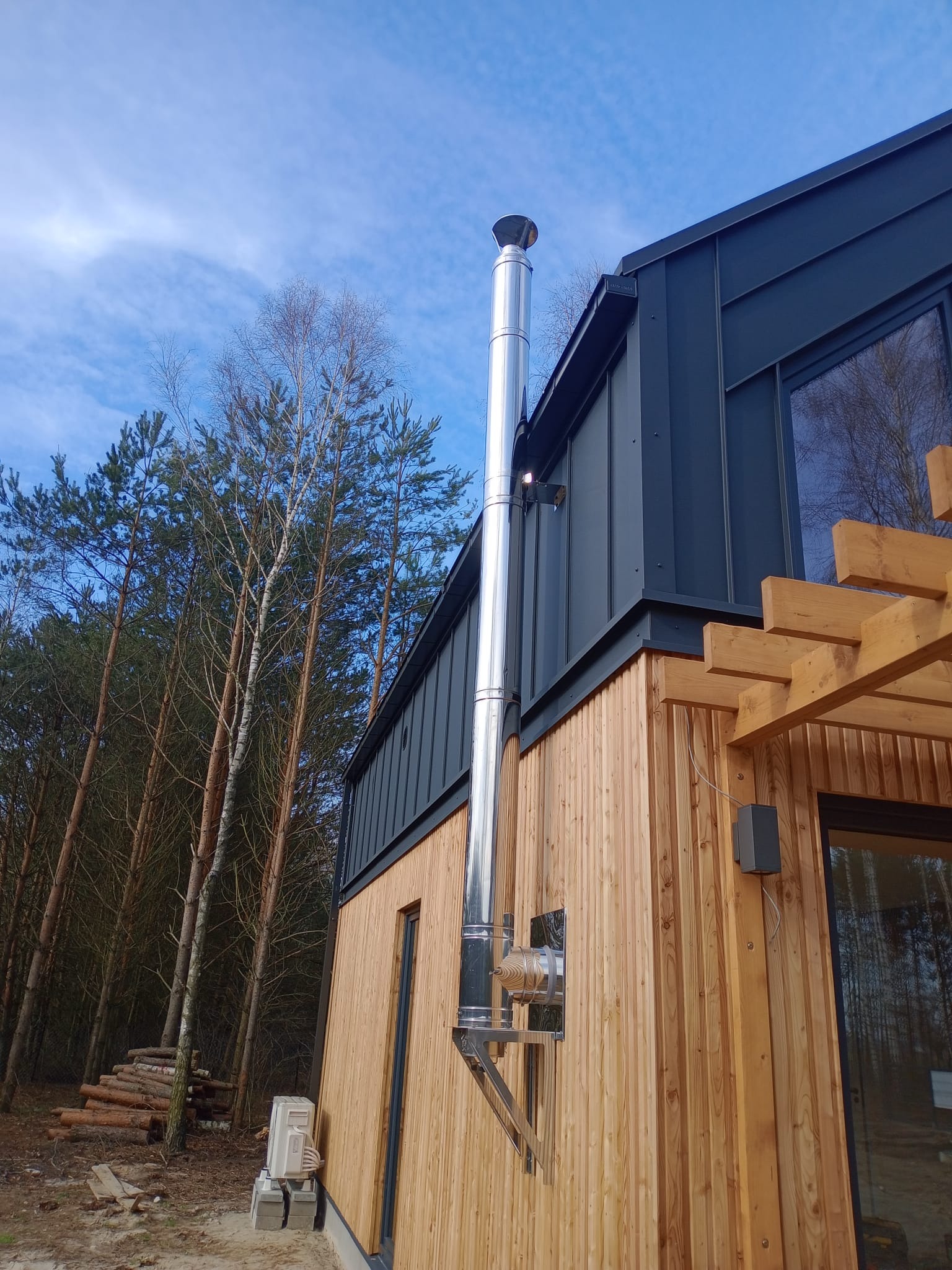 Zewnętrzny widok na fragment domu z drewnianą elewacją, ciemną blachą na dachu i kominem ze stali nierdzewnej, z widocznym stosem drewna w tle.