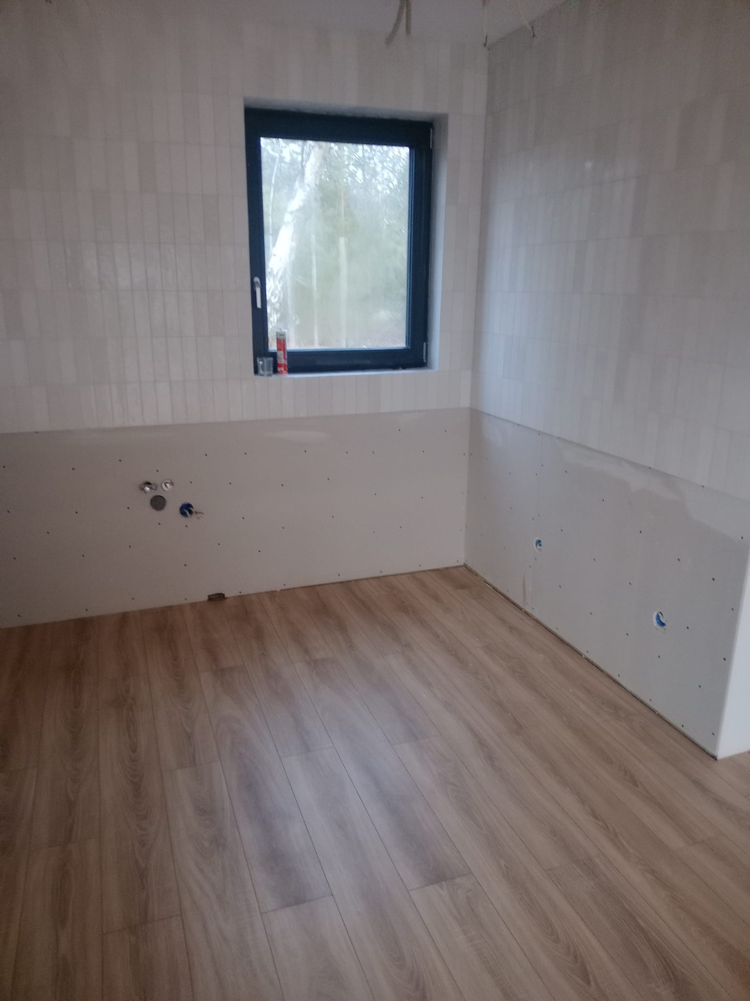 Łazienka w trakcie wykończenia: ułożone panele podłogowe, częściowo wyłożone płytki na ścianach, przygotowane przyłącza wodne i kanalizacyjne, okno z widokiem na zewnątrz.