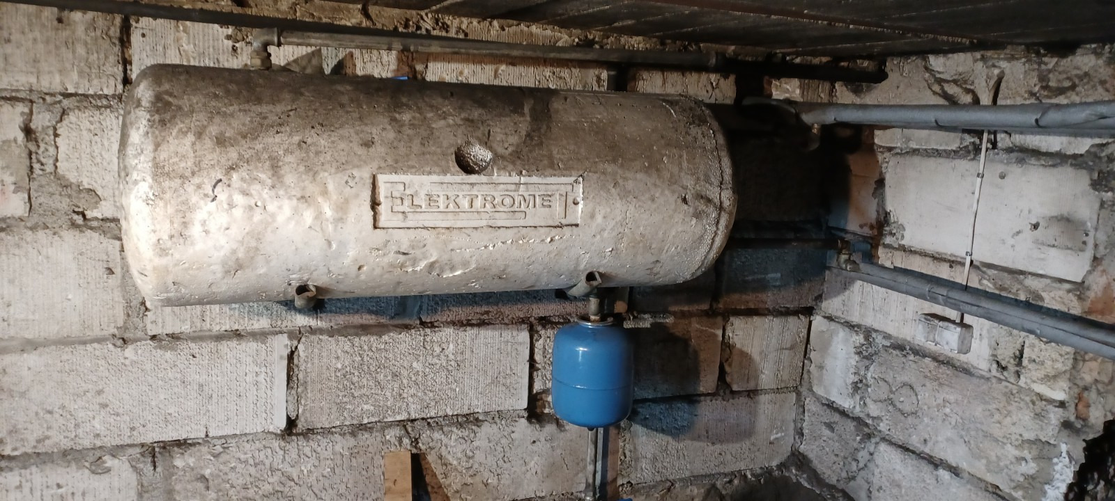 Stary, poziomy zbiornik na wodę marki Elektromet, zamontowany na ścianie z betonowych pustaków, z widocznymi rurami i niebieskim naczyniem przeponowym poniżej.