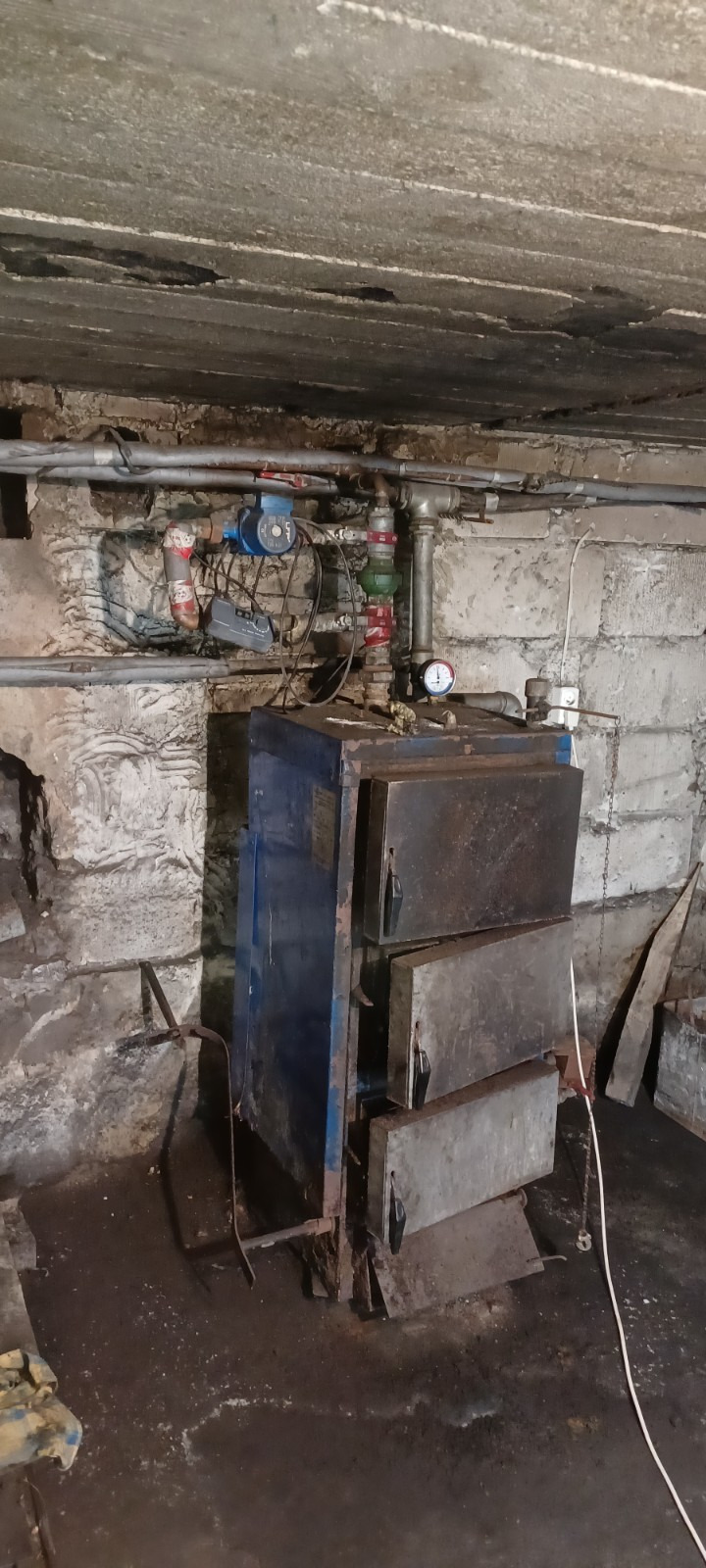 Stary, zardzewiały piec centralnego ogrzewania z trzema drzwiczkami w piwnicy z widocznymi rurami, pompą obiegową i manometrem na ścianie z kamienia.