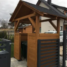 Drewniana zabudowa śmietnikowa zadaszona, z widocznymi pojemnikami na odpady, na tle domu z ciemną dachówką i szarym niebem.