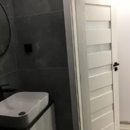 Nowoczesna łazienka z białą umywalką nablatową, ciemnoszarą płytką na ścianie i białą, częściowo przeszkloną, otwartą ościeżnicą drzwiową z czarną klamką.