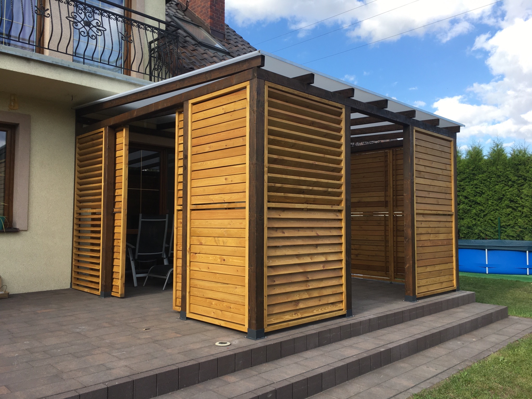Zadaszony taras z drewnianymi żaluzjami, brązowa kostka brukowa na podłodze, w tle zielony trawnik i niebieski basen.