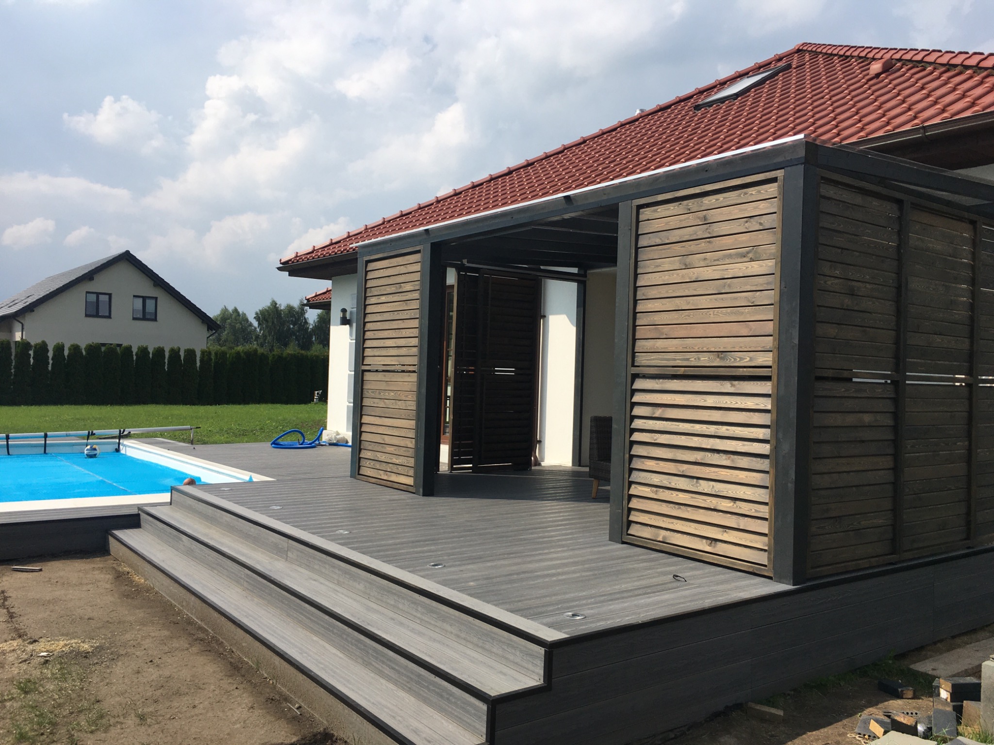 Nowoczesny taras kompozytowy z trzema schodkami prowadzącymi do basenu, z drewnianą pergolą i przesuwanymi panelami, na tle domu z czerwoną dachówką i zielonego trawnika.