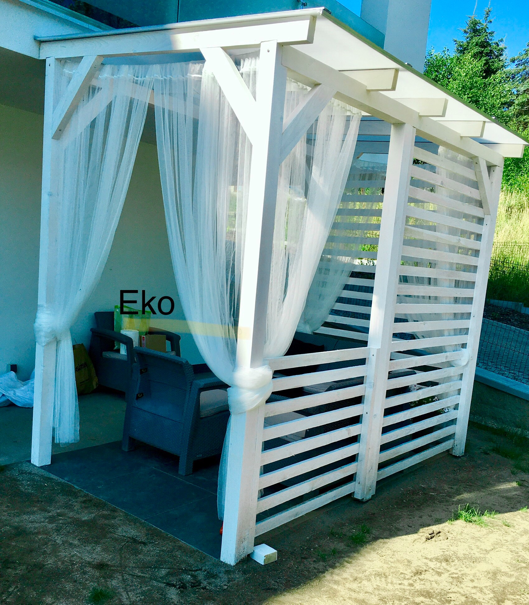 Biała, drewniana altana z ażurowymi ścianami i zwiewnymi, białymi zasłonami, z szarymi meblami ogrodowymi w środku, na zewnątrz budynku.