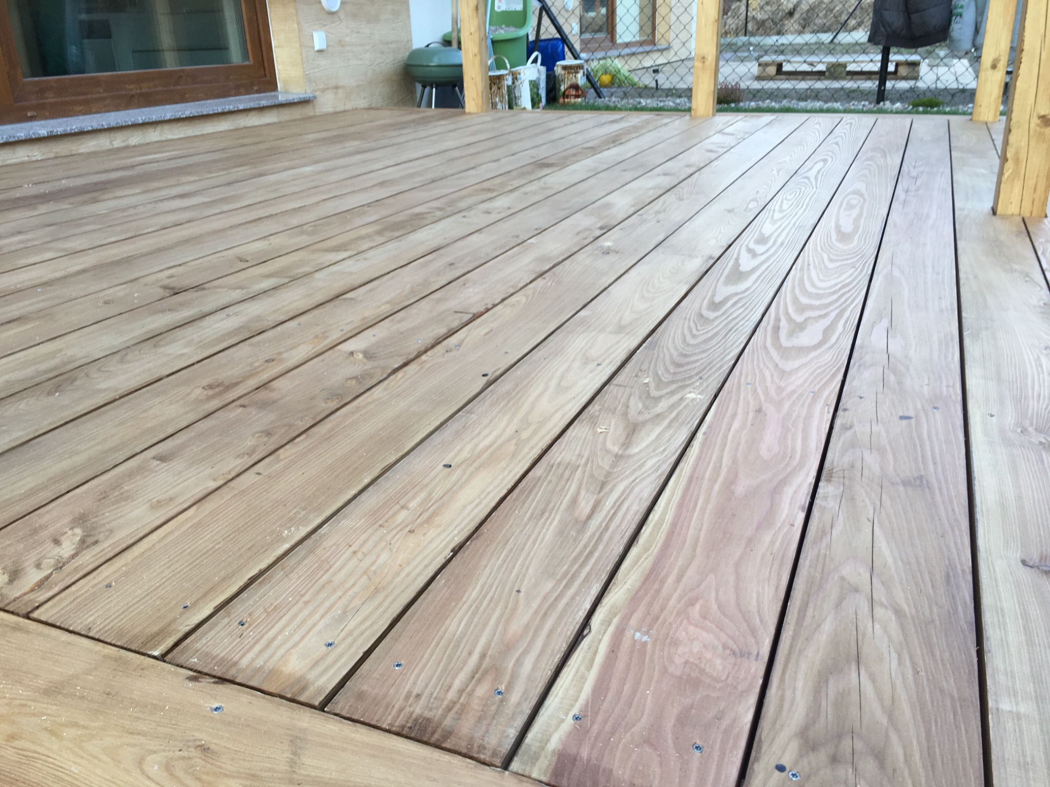 Nowo wybudowany taras z desek drewnianych, widoczne wkręty mocujące, na drugim planie grill i elementy wyposażenia ogrodu.