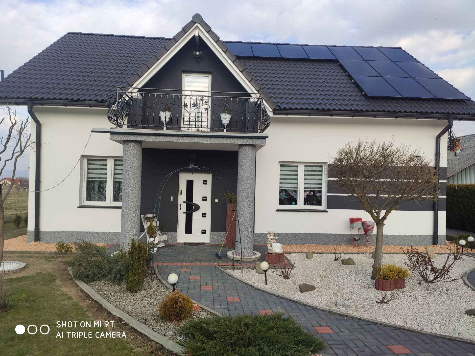 Nowoczesny dom jednorodzinny z białą elewacją, czarnym dachem z panelami słonecznymi, balkonem z ozdobną balustradą i zadbanym ogrodem z brukowanym chodnikiem.