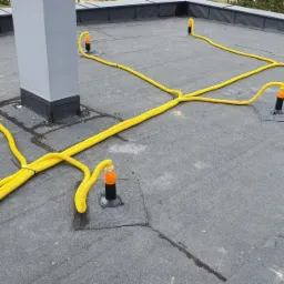 Ż&oacute;łte, karbowane rury wentylacyjne rozprowadzone po płaskim dachu z papy, zakończone pionowymi kominkami wentylacyjnymi z pomarańczowymi elementami, widoczny fragment komina wentylacyjnego.