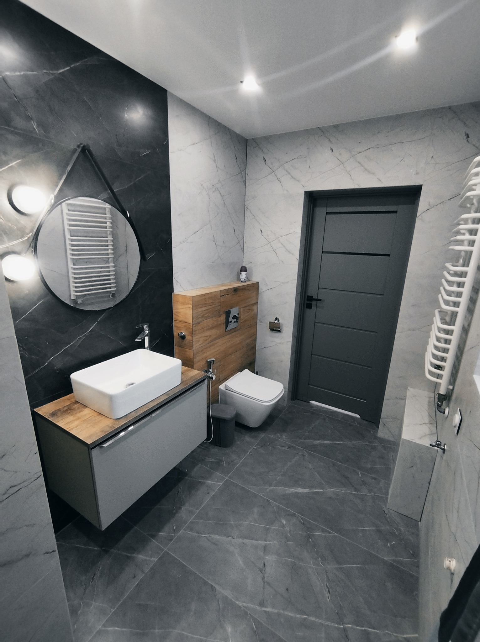 Nowoczesna łazienka z szarymi marmurkowymi płytkami, podwieszaną toaletą, umywalką nablatową na drewnianej szafce i okrągłym lustrem z oświetleniem LED.