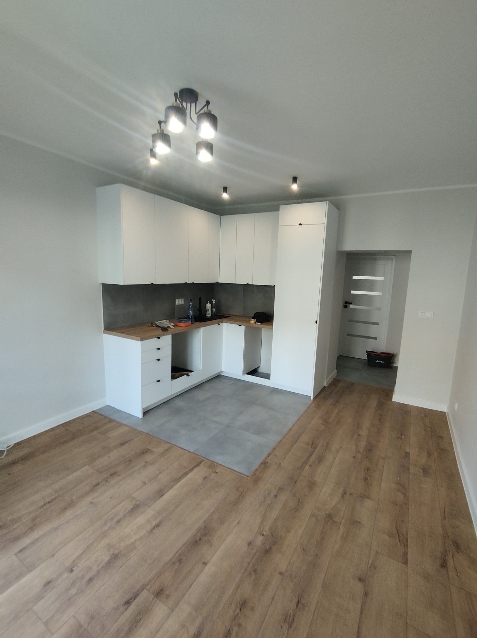 Wnętrze kuchni w trakcie montażu: białe szafki kuchenne bez frontów, szara ściana nad blatem, drewnopodobne panele podłogowe, widoczna instalacja elektryczna i hydrauliczna.