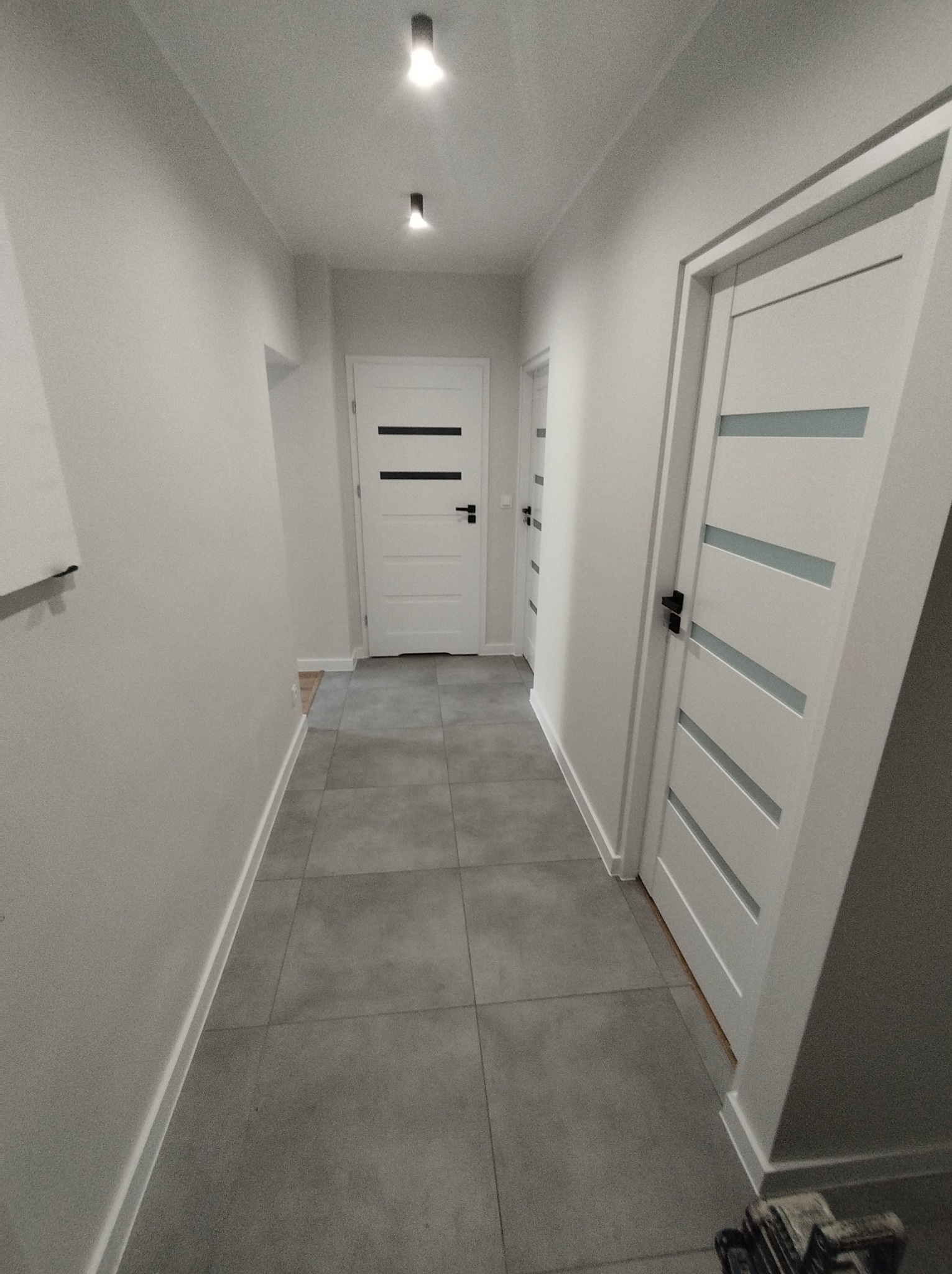 Nowoczesny korytarz w odcieniach szarości i bieli, z szarymi płytkami podłogowymi i białymi drzwiami z poziomymi przeszkleniami, oświetlony punktowymi lampami sufitowymi.