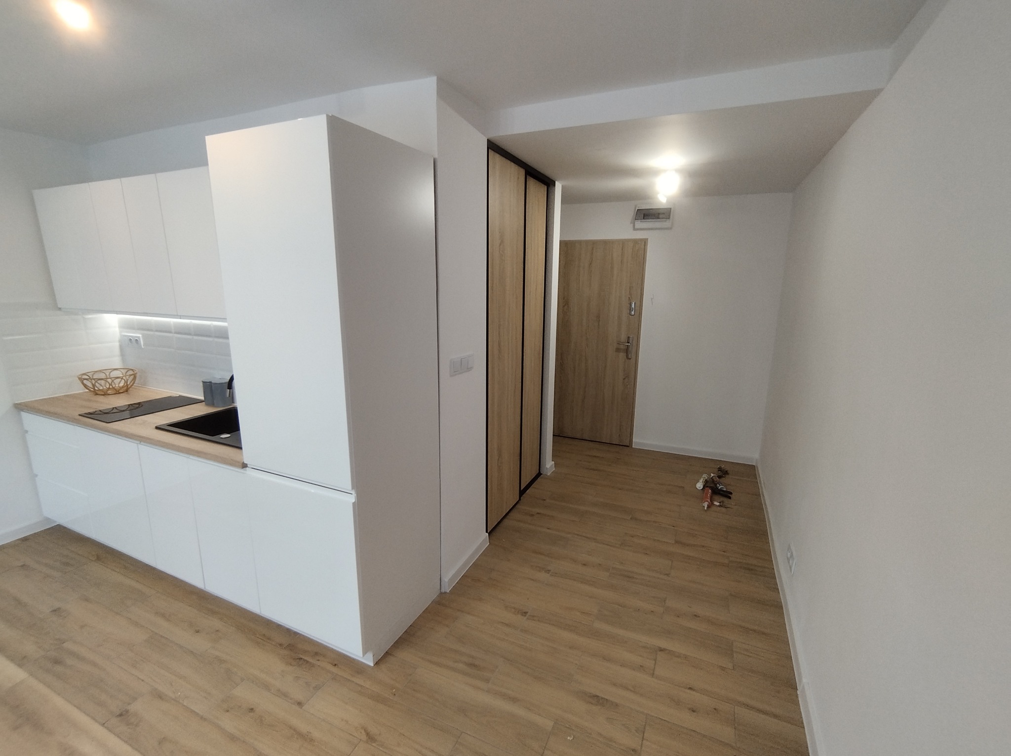 Nowoczesna kuchnia z białymi, lakierowanymi frontami szafek, drewnianym blatem i czarnym zlewem, widok na przedpokój z drewnianymi drzwiami i rozrzuconymi narzędziami budowlanymi na podłodze.