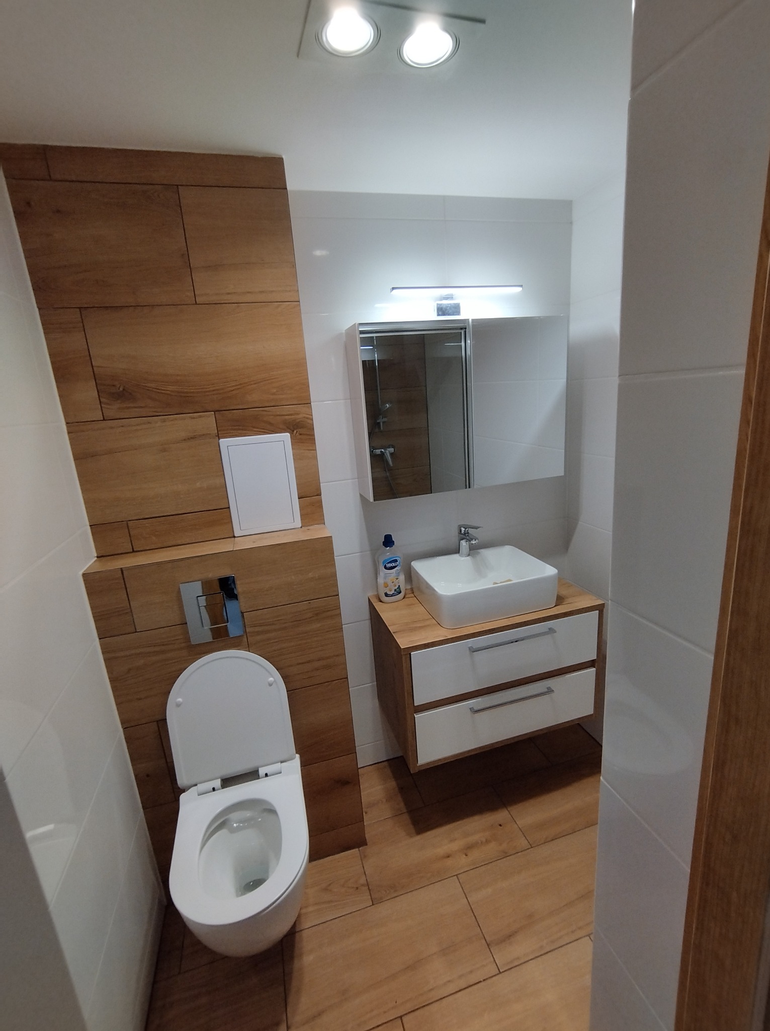 Nowoczesna łazienka z drewnopodobnymi płytkami na ścianie i podłodze, podwieszana toaleta z ukrytym systemem spłukiwania, biała umywalka nablatowa z szafką i lustrem z oświetleniem LED.