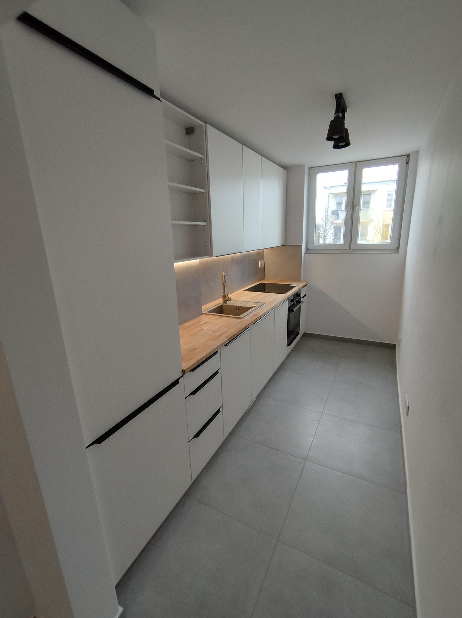 Minimalistyczna kuchnia w odcieniach bieli i szarości z drewnianym blatem, oświetleniem LED pod szafkami wiszącymi i czarnymi, podłużnymi uchwytami szafek dolnych.