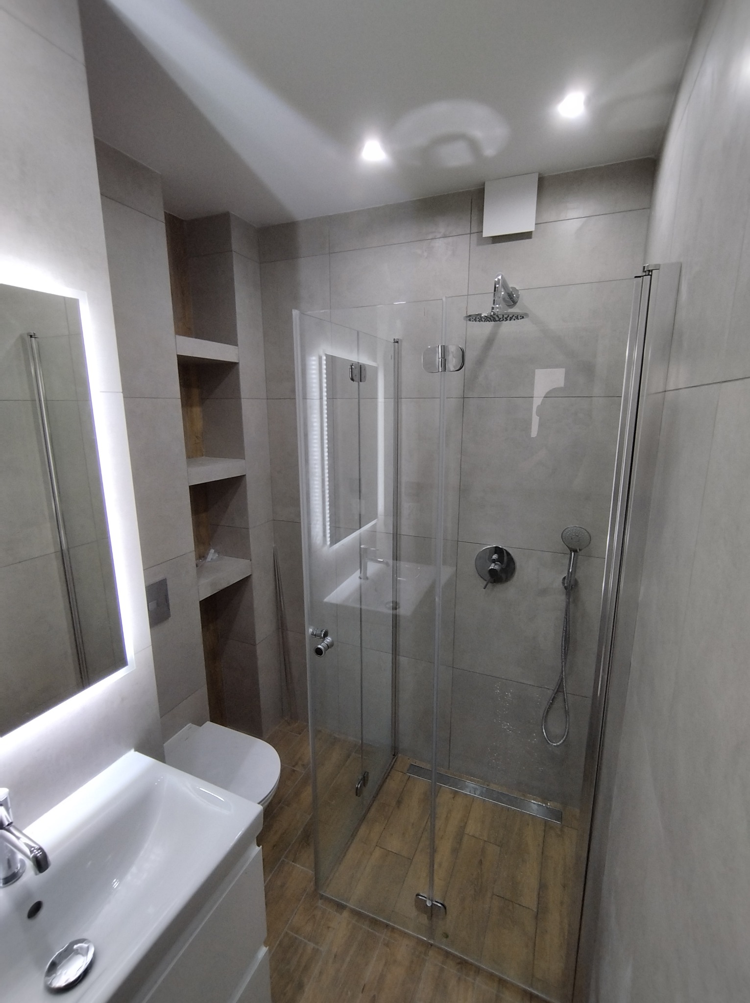 Nowoczesna łazienka z kabiną prysznicową typu walk-in, oświetlonym lustrem i wnęką z półkami, wykończona płytkami w odcieniach szarości i podłogą imitującą drewno.