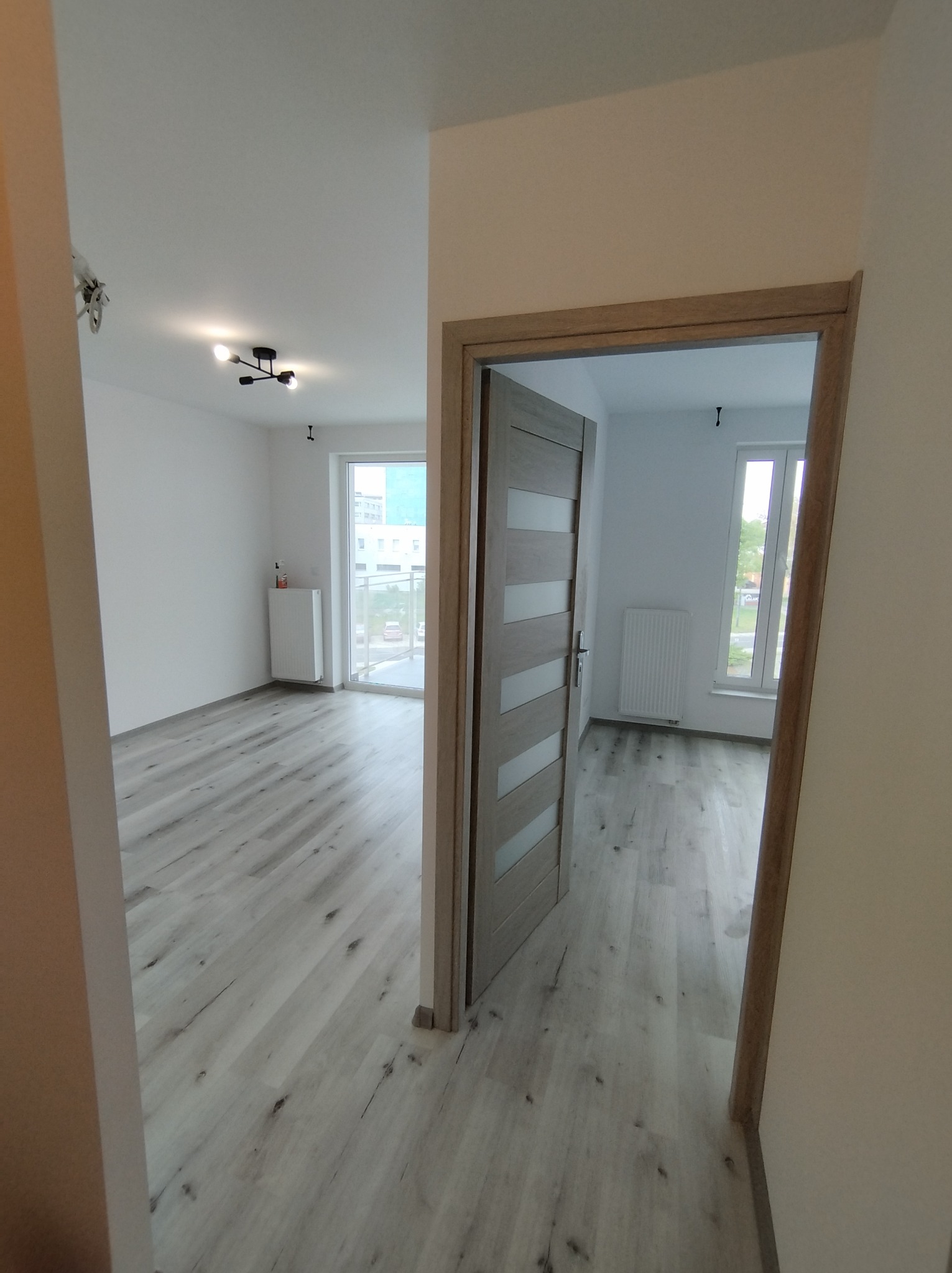 Widok przez otwarte drzwi na świeżo wykończone, puste wnętrze mieszkania z panelami podłogowymi imitującymi drewno, białymi ścianami, grzejnikiem i nowoczesną lampą sufitową.