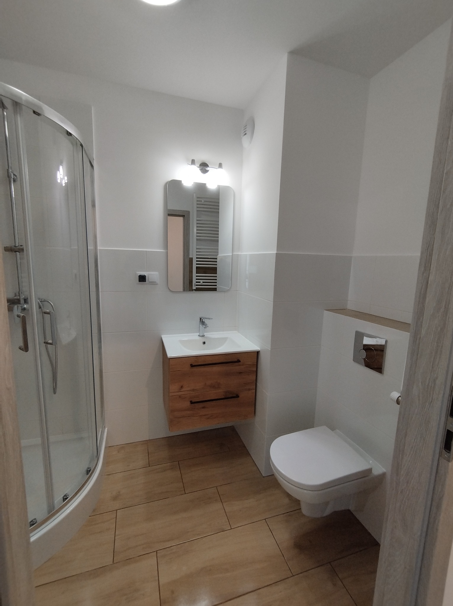 Nowoczesna, jasna łazienka z kabiną prysznicową z zaokrąglonym frontem, szafką pod umywalką z drewnianymi szufladami i wiszącą toaletą, wykończona płytkami w odcieniach bieli i beżu.