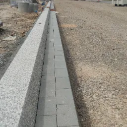 Ułożone obrzeże chodnika z szarej kostki betonowej, przylegające do granitowego krawężnika w trakcie budowy drogi, widoczne maszyny budowlane w tle.