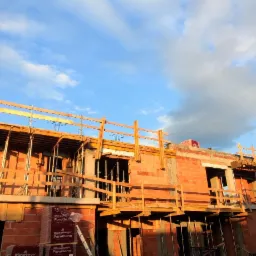 Surowy, dwukondygnacyjny dom murowany w trakcie budowy, z widocznymi czerwonymi cegłami Porotherm, drewnianym szalunkiem i małą choinką na dachu w tle błękitnego, pochmurnego nieba.