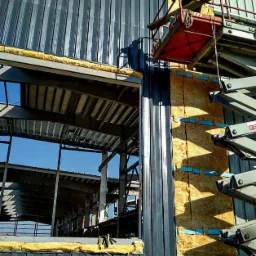 Montaż płyt warstwowych na hali stalowej z użyciem podnośnika, widoczna izolacja termiczna i stalowa konstrukcja wewnątrz.