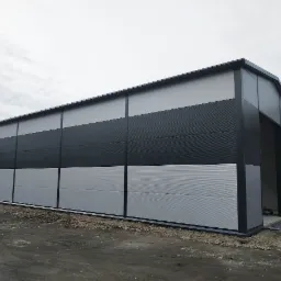 Gotowa hala przemysłowa z paneli warstwowych w odcieniach szarości, widoczna boczna ściana z dużym otworem wjazdowym, teren wokół jeszcze niezagospodarowany.