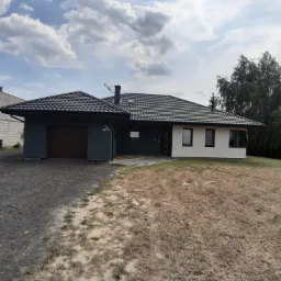 Jednopiętrowy dom z garażem i ciemną dachówką, część elewacji w kolorze grafitowym, druga część biała, numer domu 7A.