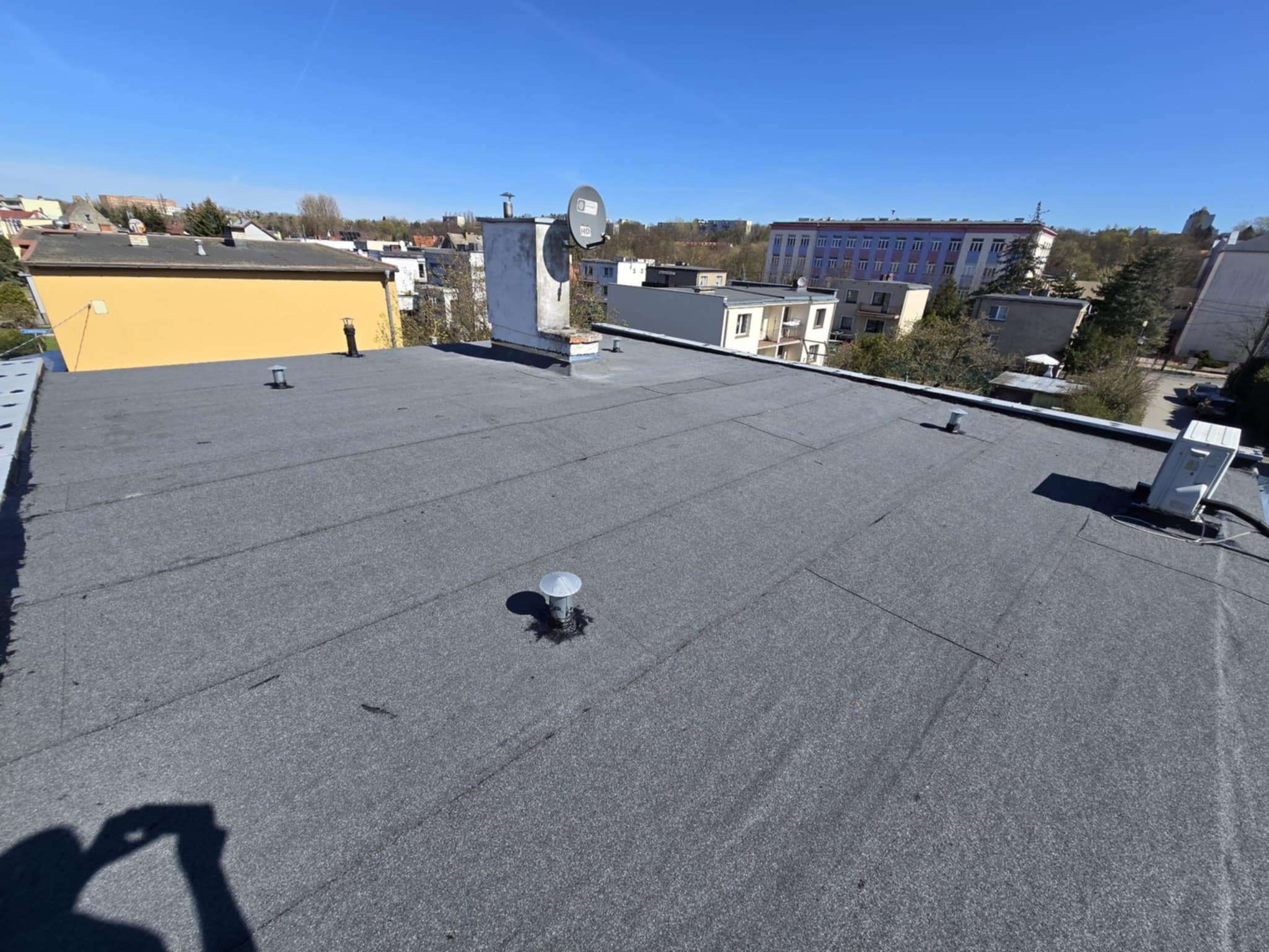 Szary, płaski dach pokryty papą na tle zabudowy miejskiej w słoneczny dzień. Widoczne kominy, antena satelitarna i elementy wentylacji.